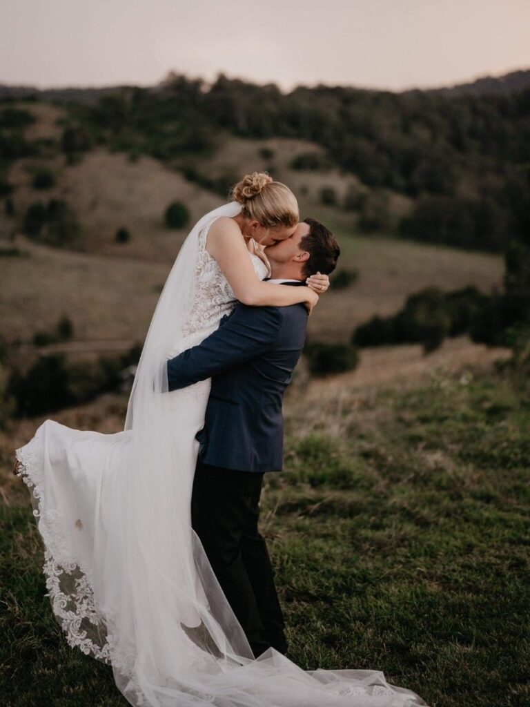 The bride, in an elegant white lace dress, is lifted by her groom in a tender kiss. Behind them, rolling countryside stretches into the distance, capturing the moment.